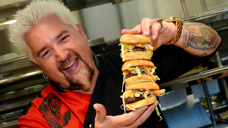Guy Fieri laughing and holding three burgers stacked together