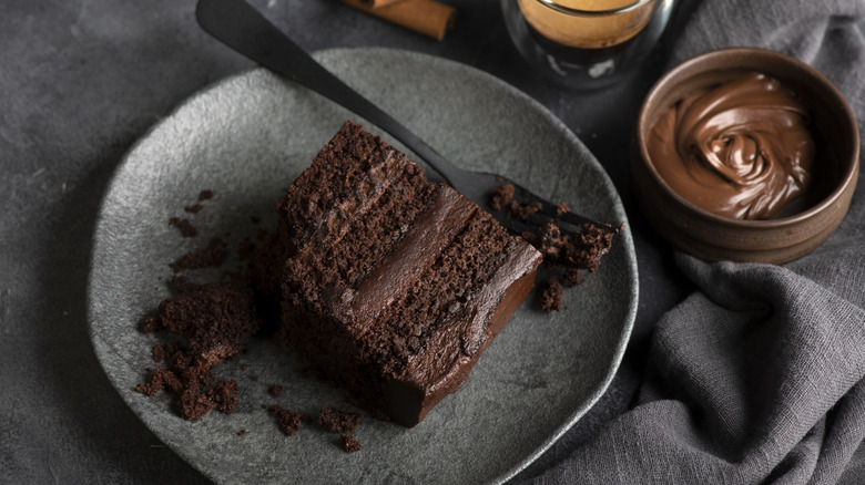 A slice of chocolate cake sits on a plate next to a small bowl of frosting.