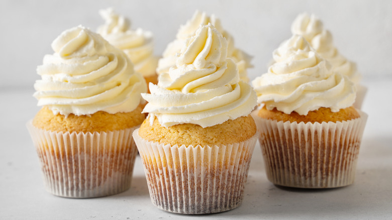 Three vanilla cupcakes with vanilla frosting are on a table.