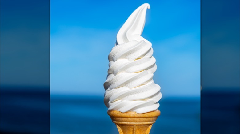 A vanilla soft serve ice cream in a cone is set against a blue sky.