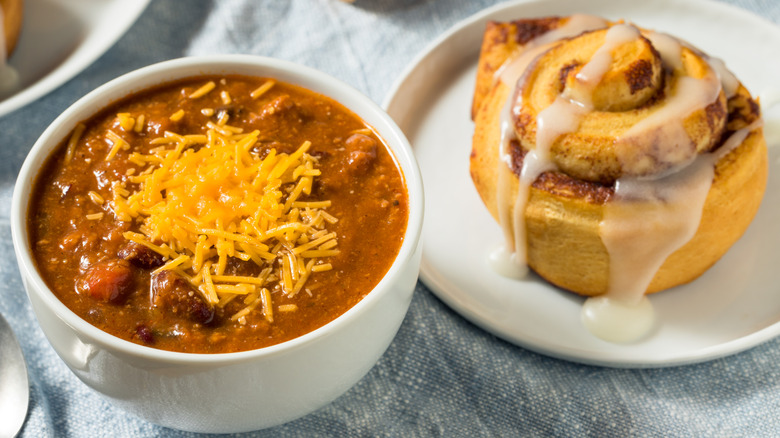 a white bowl of chili with cheese and a plate with a cinnamon roll beside it.