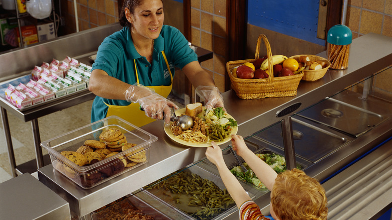 a cafeteria worked scoops meatballs onto a yellow plate full of food for a young student.