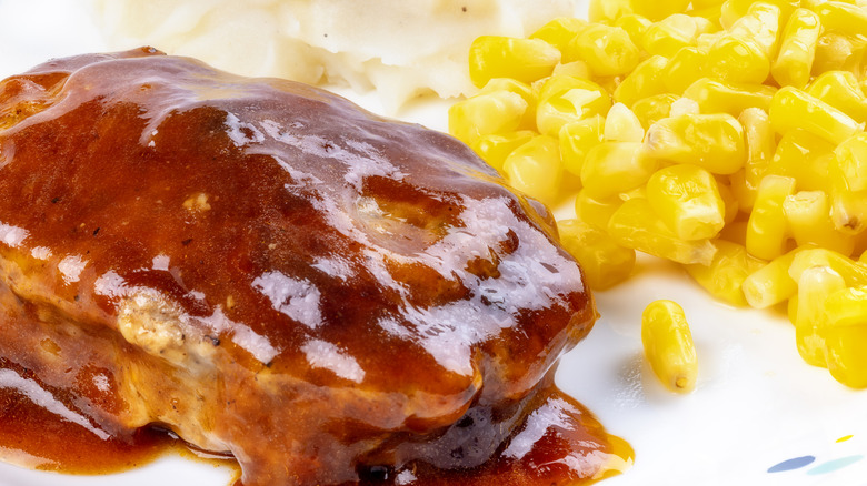 a loaf of meat covered in a shiny brown sauce beside corn kernels and mashed potatoes