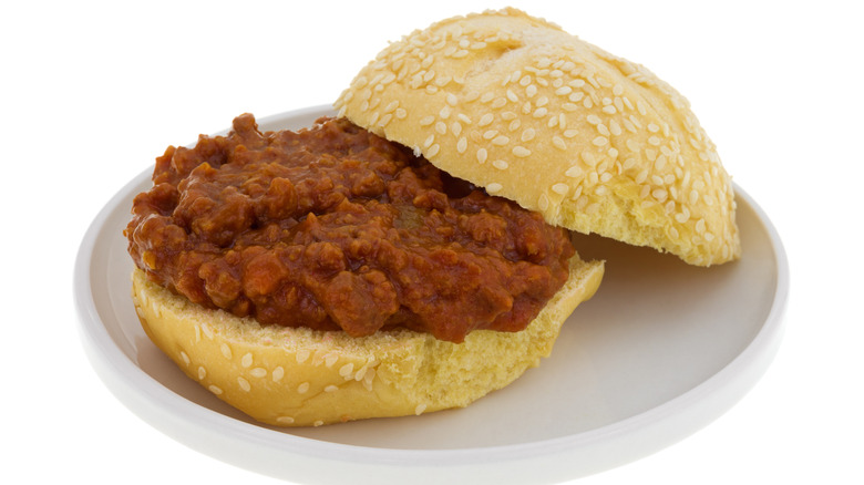 a sesame seed hamburger bun with sloppy joe filling on a white circle plate.