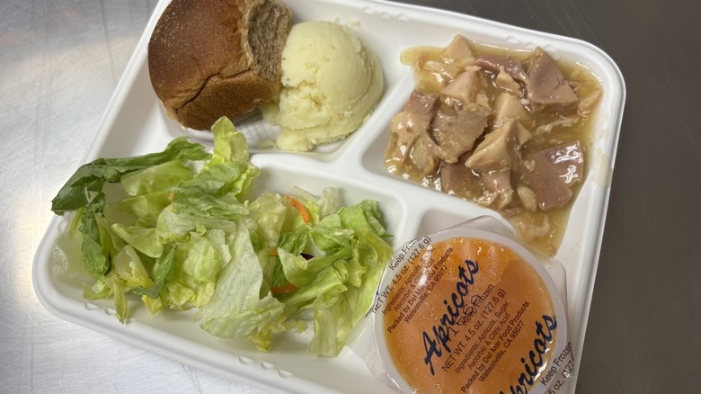 a white lunch tray with turkey in gravy, a scoop of mashed potatoes, a roll, salad, and cup of fruit.