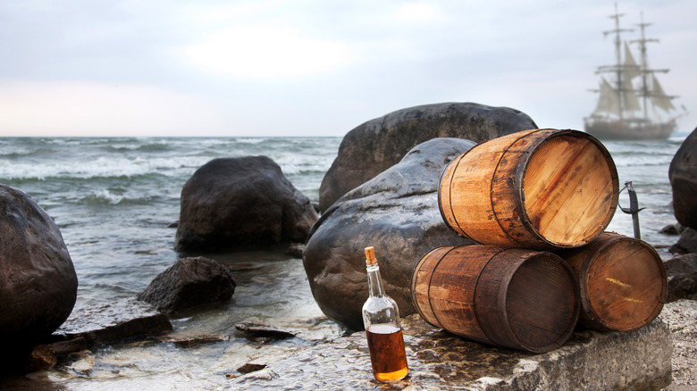 A bottle of rum on the beach next to three casks and a sword with a ship in the background.