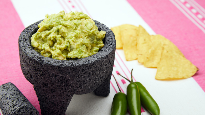 Guacamole in a molcajete with chips and jalapeños on a white and pink tablecloth.