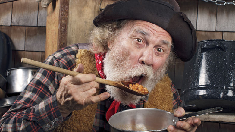 An older man dressed as a prospector is eating beans from a pot.
