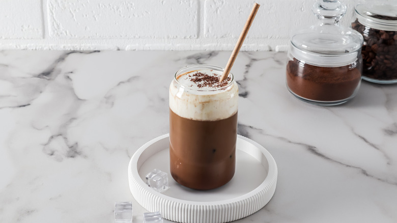 an iced coffee drink topped with creamy white foam in a glass standing on a coaster on a marble table