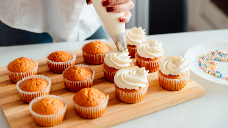 woman's hand piping frosting onto a board full of vanilla cupcakes next to a plate of sprinkles
