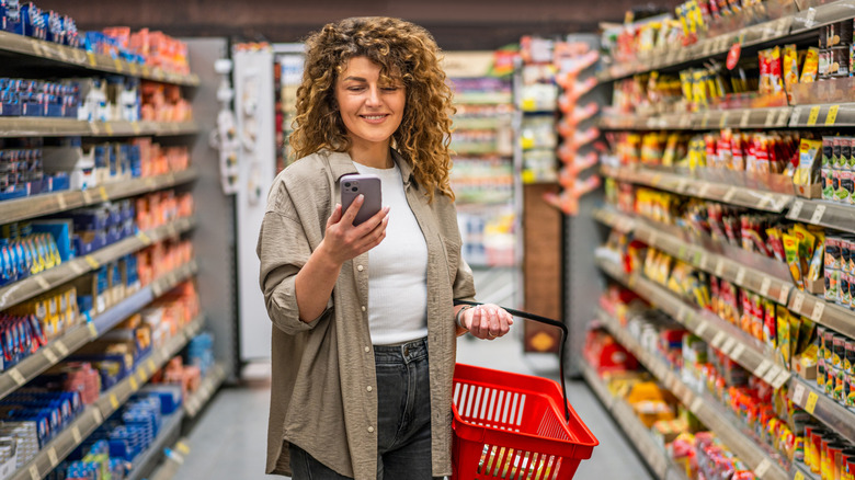 Woman stands in grocery store aisle holding a shopping basket in one hand, looking at the phone in her other