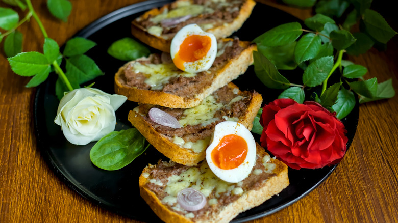 Bulgarian Princess toast with meat, cheese, onion, and hard-boiled eggs on a black plate adorned with a white and red rose