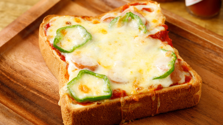 Slice of pizza toast on thick bread with cheese and green bell pepper rings on a wooden board