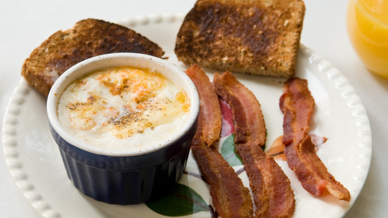 Shirred eggs in a dark blue ramekin with toast and bacon on plate