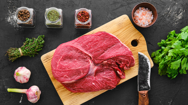 Beef top round roast on a wooden cutting board surrounded with spices in jars, fresh thyme, parsley, and heads of garlic