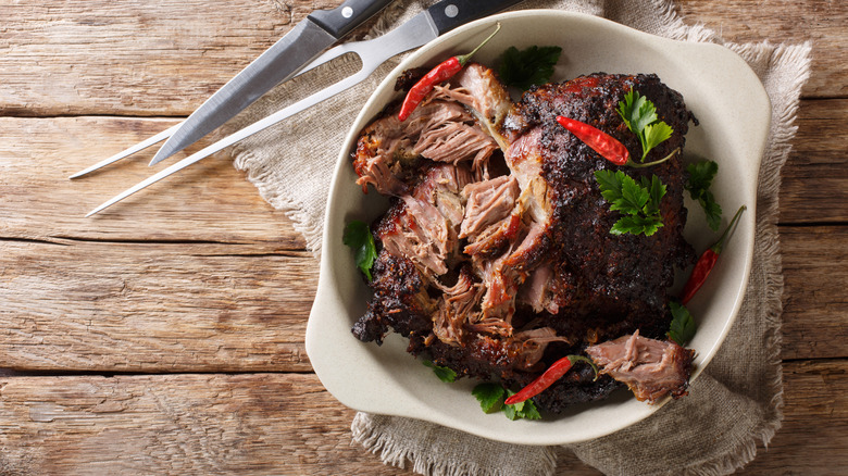 Puerto Rican pérnil in a white bowl topped with chilis and parsley with carving utensils alongside the dish