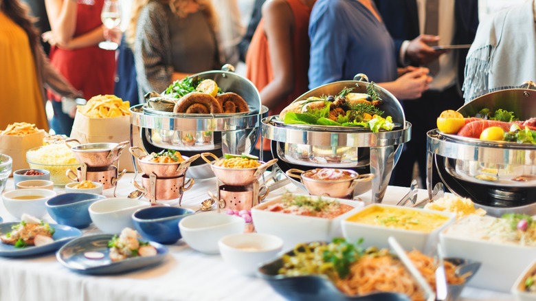 A buffet with large silver serving dishes and people in the background