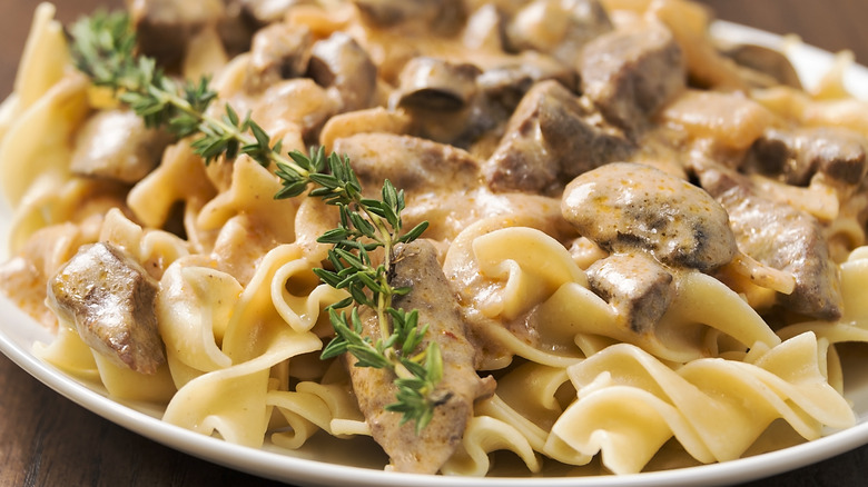 Closeup shot of creamy beef stroganoff atop egg noodles with a stem of fresh thyme laid atop it as garnish