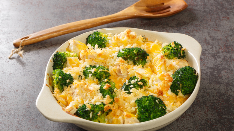 Oval casserole dish filled with chicken divan, which is a casserole of chicken, rice, and broccoli, and a wooden spoon lies behind it on the counter