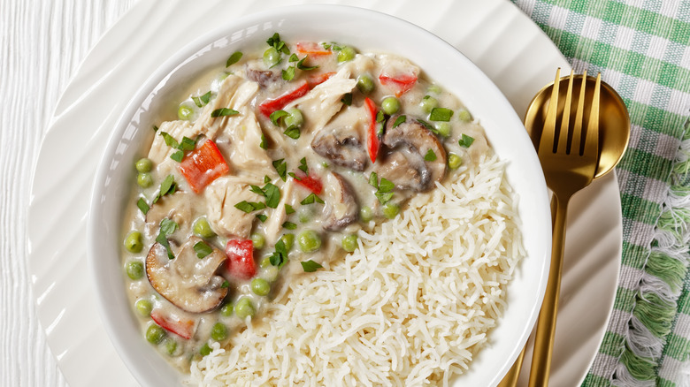 View looking straight down of a white bowl filled with chicken à la king, with mushrooms and red peppers, with rice on part of the plate and utensils beside it