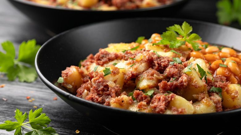 Closeup shot of corned beef hash on a black plate with beans behind it, topped with parsley garnish