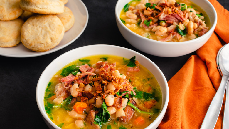 Two bowls of ham and bean soup (in foreground and background) topped with bacon crumbles sitting on a table with a plate of biscuits in the background