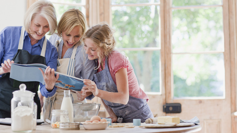 Three generations of women in aprons stand in front of a kitchen counter looking at the same book with baking ingredients spread out on the counter before them