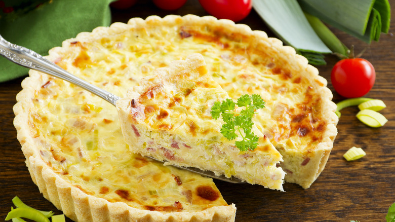 Whole quiche Lorraine with a triangular slice of quiche cut out and being lifted up with a spatula