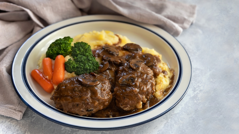 Plate with Salisbury steak topped with mushrooms and brown gravy with mashed potatoes, carrots, and broccoli behind it on the plate