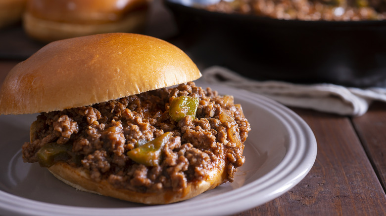 Sloppy joe sandwich on a brioche bun is plated on a wooden table with the cast iron skillet full of sloppy joe meat in the background