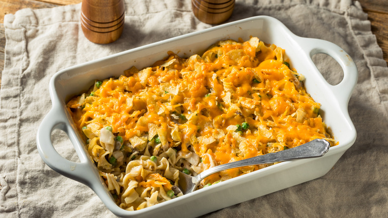 Rectangular casserole dish filled with tuna noodle casserole topped with cheese and crumbled potato chips, with a spoon scooping out a corner of the casserole