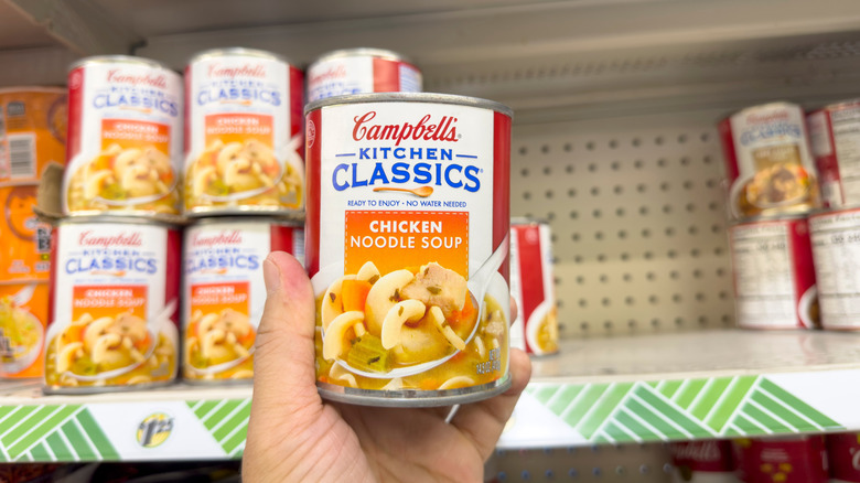 A hand holding a can of Campbell's Kitchen Classic Chicken Noodle Soup, at the store