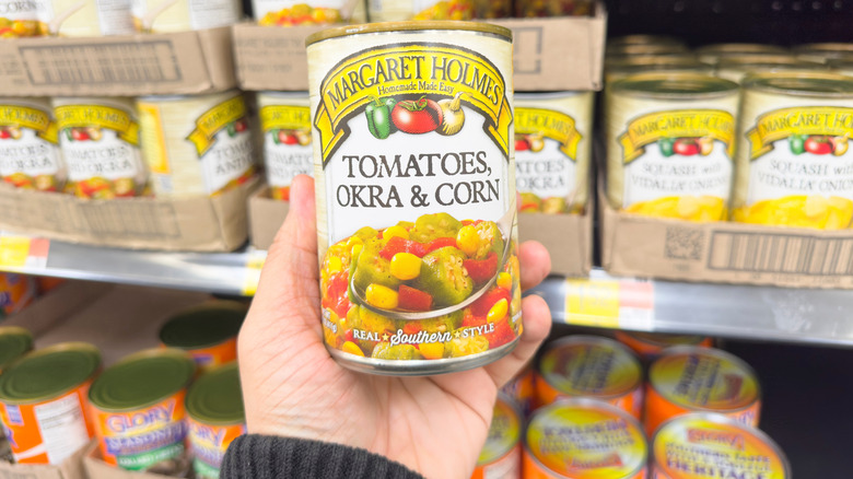A hand holding a can of Margaret Holmes Tomatoes, Okra & Corn at a grocery store