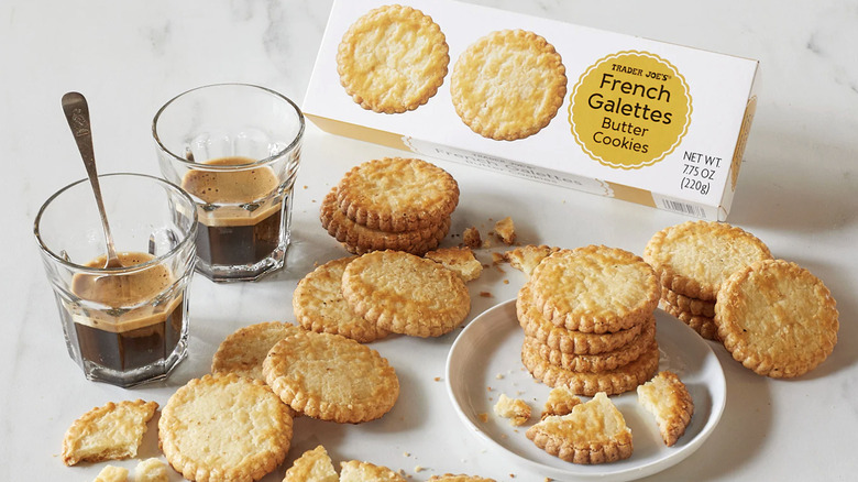 several butter cookies on a plate and table next to two cups of coffee with box of trader joe's cookies behind them