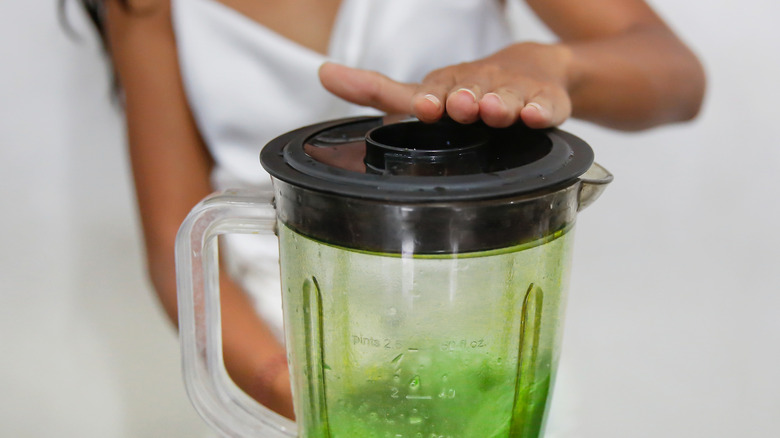 a person's hand on a blender lid with green contents inside.
