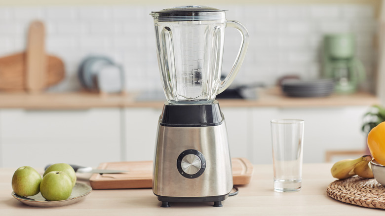 an empty glass blender on a wooden counter with produce, a cutting board, and drinking glass.
