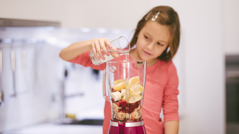 a young girl adding more liquid to a blender jar full of various fruits.