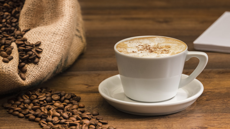 Cup of frothy cappuccino served beside an overflowing bag of coffee beans