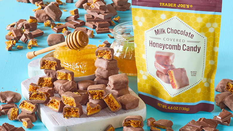 A bag of Trader Joe's Milk Chocolate Covered Honeycomb Candy sits beside many pieces of the candy as well as honeycomb and a honey dipper