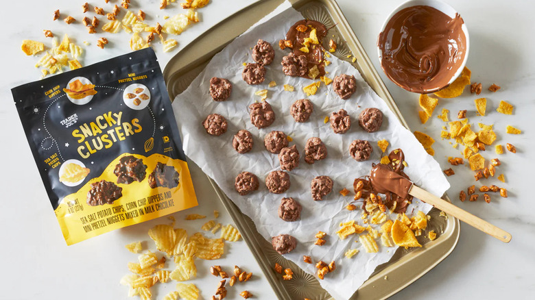 A bag of Trader Joe's Snacky Clusters sits near a pan of the treats, a bowl of melted chocolate, and crumbled snack bits