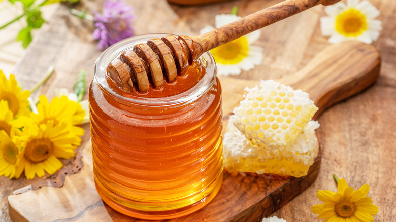 a jar of honey with a honey stick on top, flowers and honeycomb