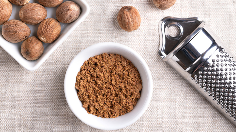 A bowl of ground nutmeg with nutmeg fruits and a grater in the background