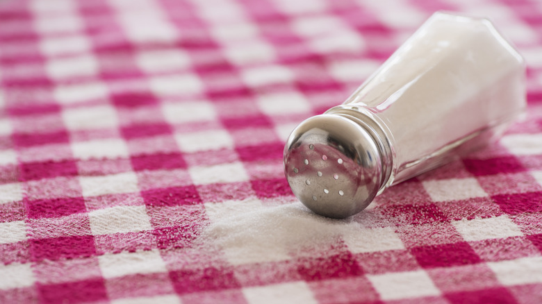 A salt shaker on its side, salt is spilled on checker tablecloth
