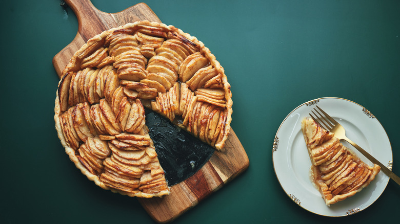 an apple pie with a slice cut out of it sits on a wooden serving board, slice on plate