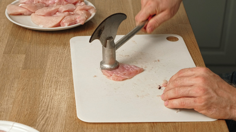 Hands pounding raw chicken on cutting board with mallet