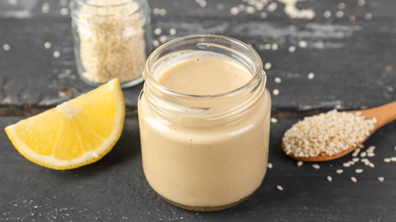 Jar of tahini between a spoonful of sesame seeds and a lemon wedge