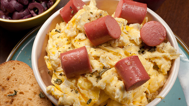 White bowl of scrambled eggs topped with chunks of hot dog
