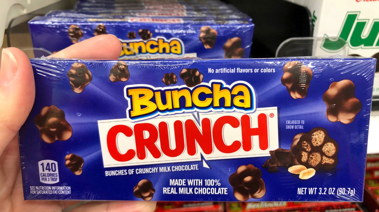 A hand holds up a package of Nestle Buncha Crunch in front of a store candy shelf