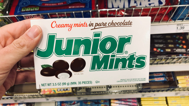 A hand holds up a box of Junior Mints in a store candy aisle