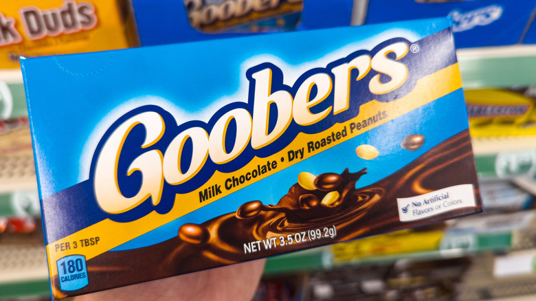 A hand holds up a box of Goobers in front of a store candy shelf